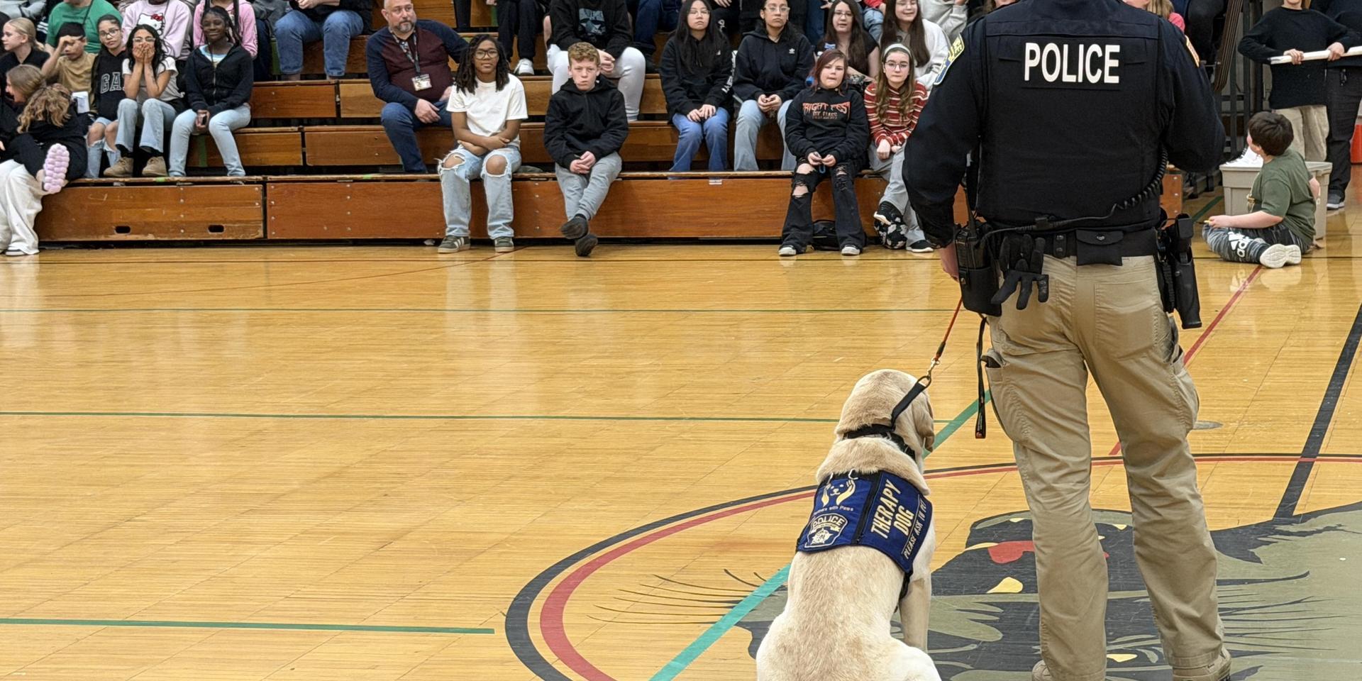 A police officer with a service dog addresses a gathering of students in a gymnasium.