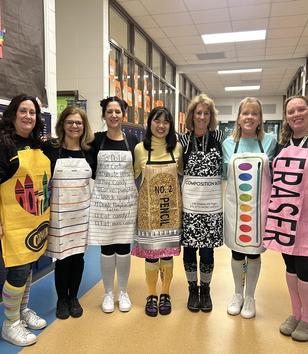 Seven women dress in creative art-themed aprons, standing together in a school hallway.