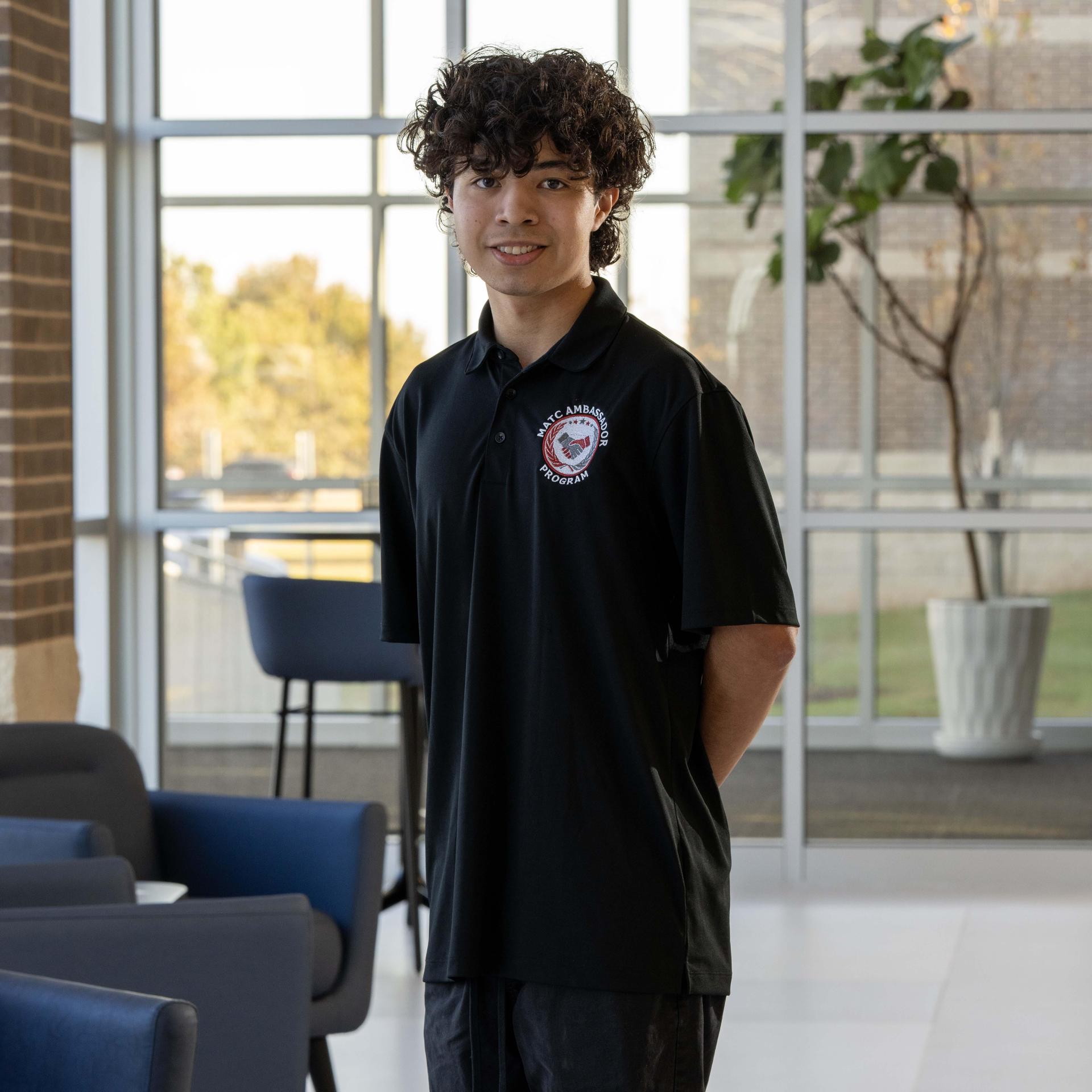Smiling young man in a black polo shirt stands in a modern indoor setting.
