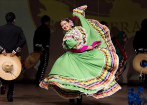 A folklorico dancer in a bright green dress and pink gloves twirls energetically on stage, smiling toward the camera as male performers in black mariachi-style outfits hold hats in the background.
