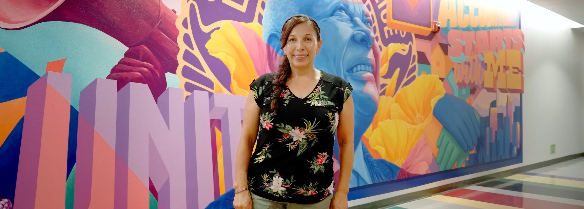 A woman stands in front of a vibrant mural with floral and colorful designs.