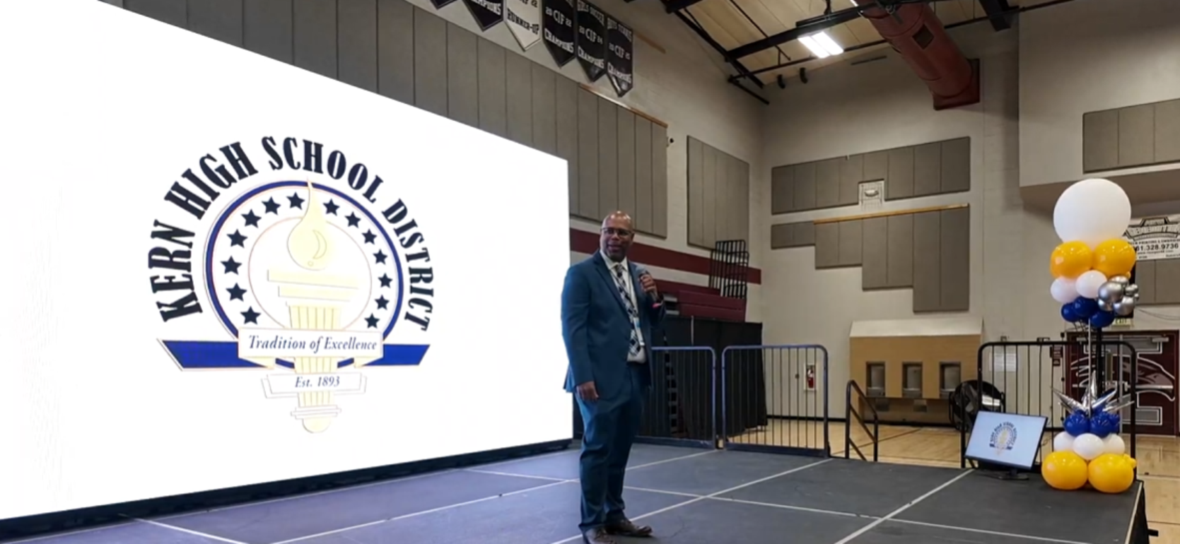 A speaker stands on stage in front of a large school district logo and decorations.