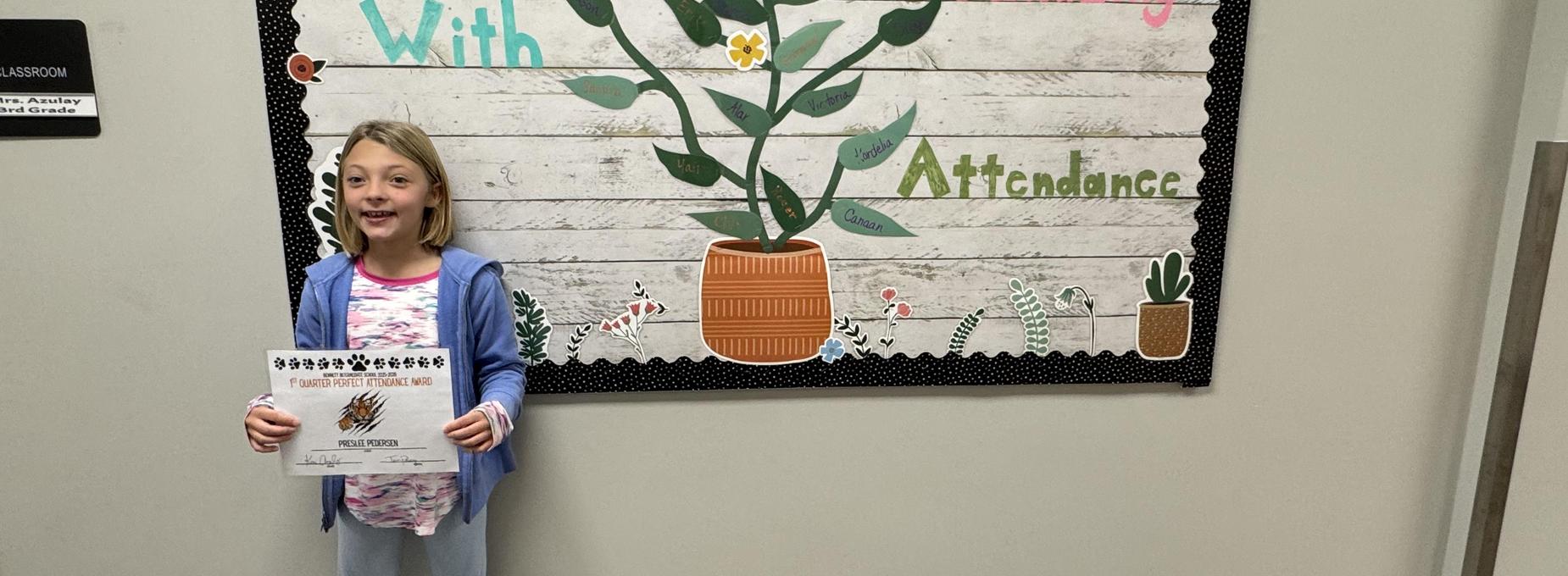 Child with a certificate standing by a plant-themed classroom banner.