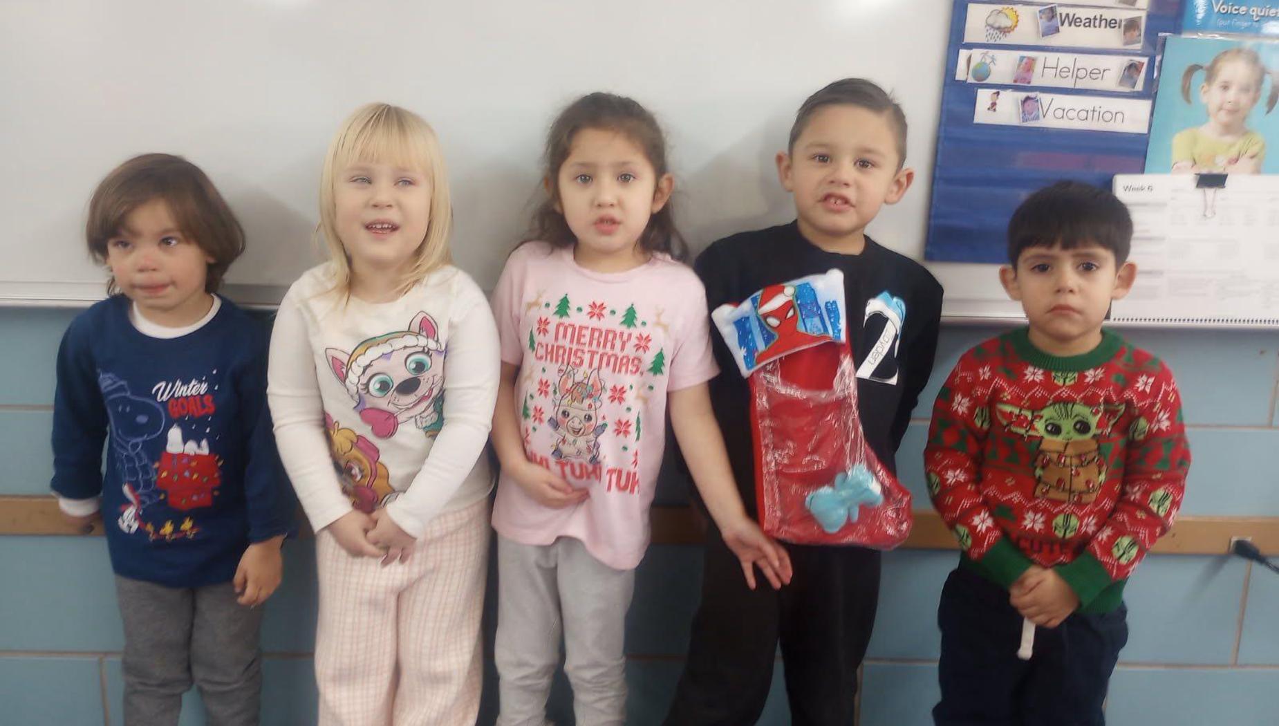 Five children in pajamas with holiday themes and a stocking in a classroom