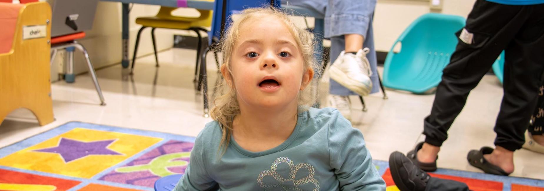 Young girl with Down syndrome sitting on a colorful classroom rug.