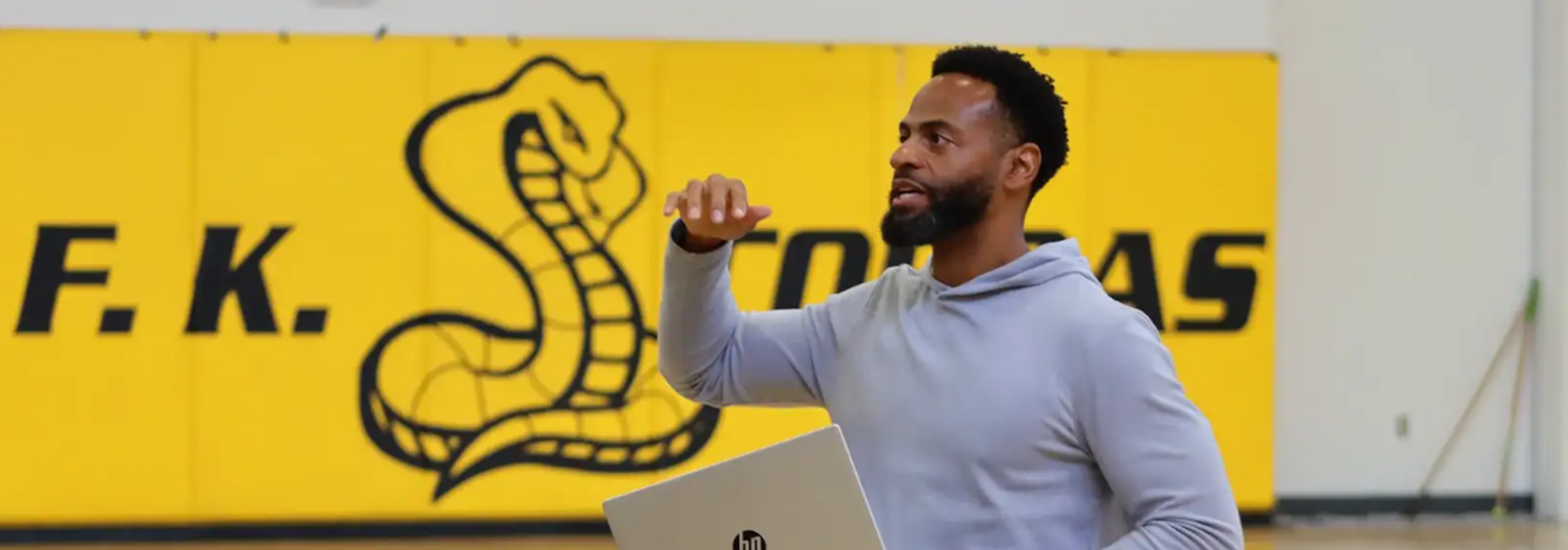 A man gesturing while holding a laptop in a gym with a yellow snake logo backdrop.