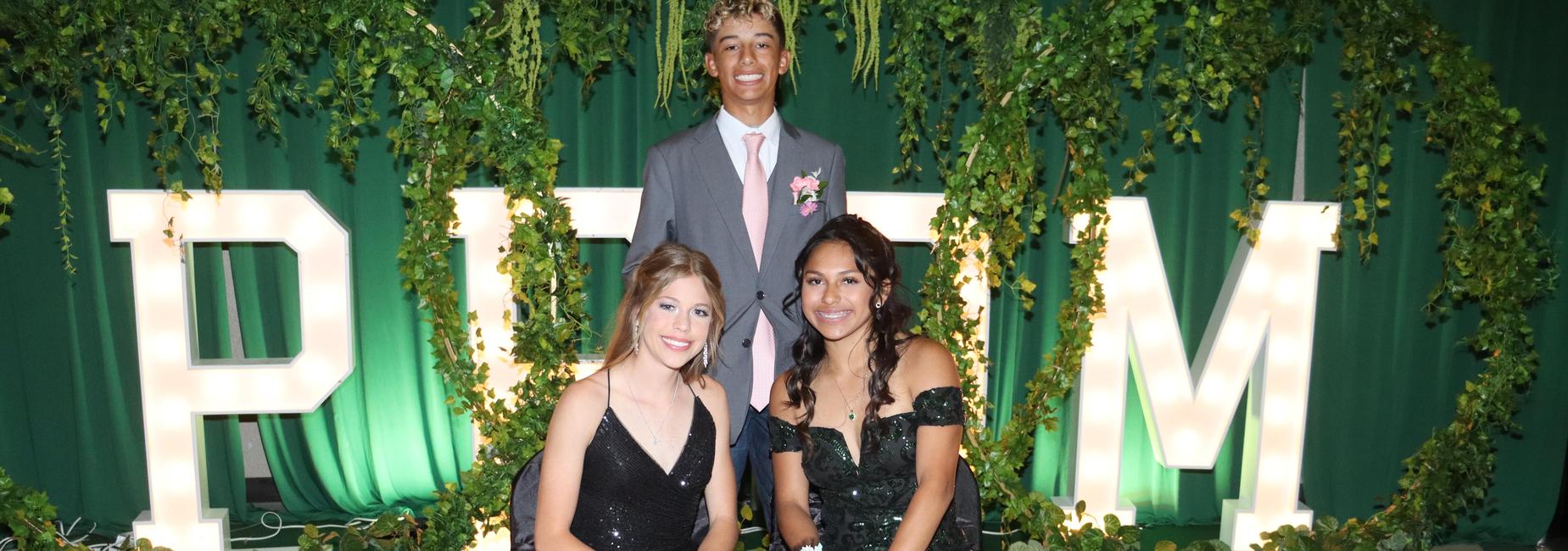 Two young women in elegant dresses sit in front of large letters, showcasing a celebratory theme.