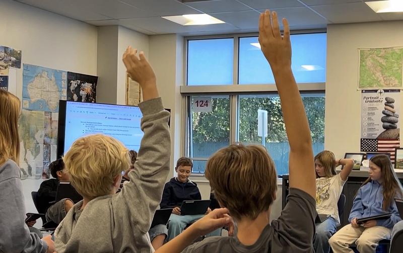 Two students raise their hands during a lesson in a classroom with a presentation displayed behind them.