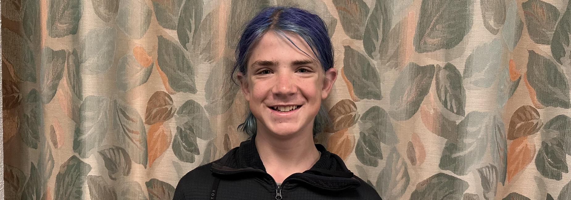 Smiling student with blue hair holding a certificate in front of a leafy curtain.