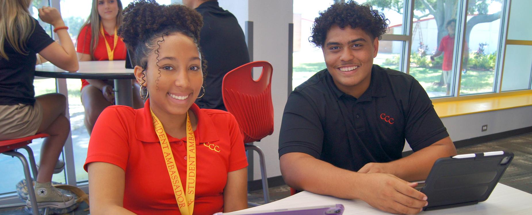 a girl in a red CCC polo and a boy in a black CCC polo look up from their iPads with smiles