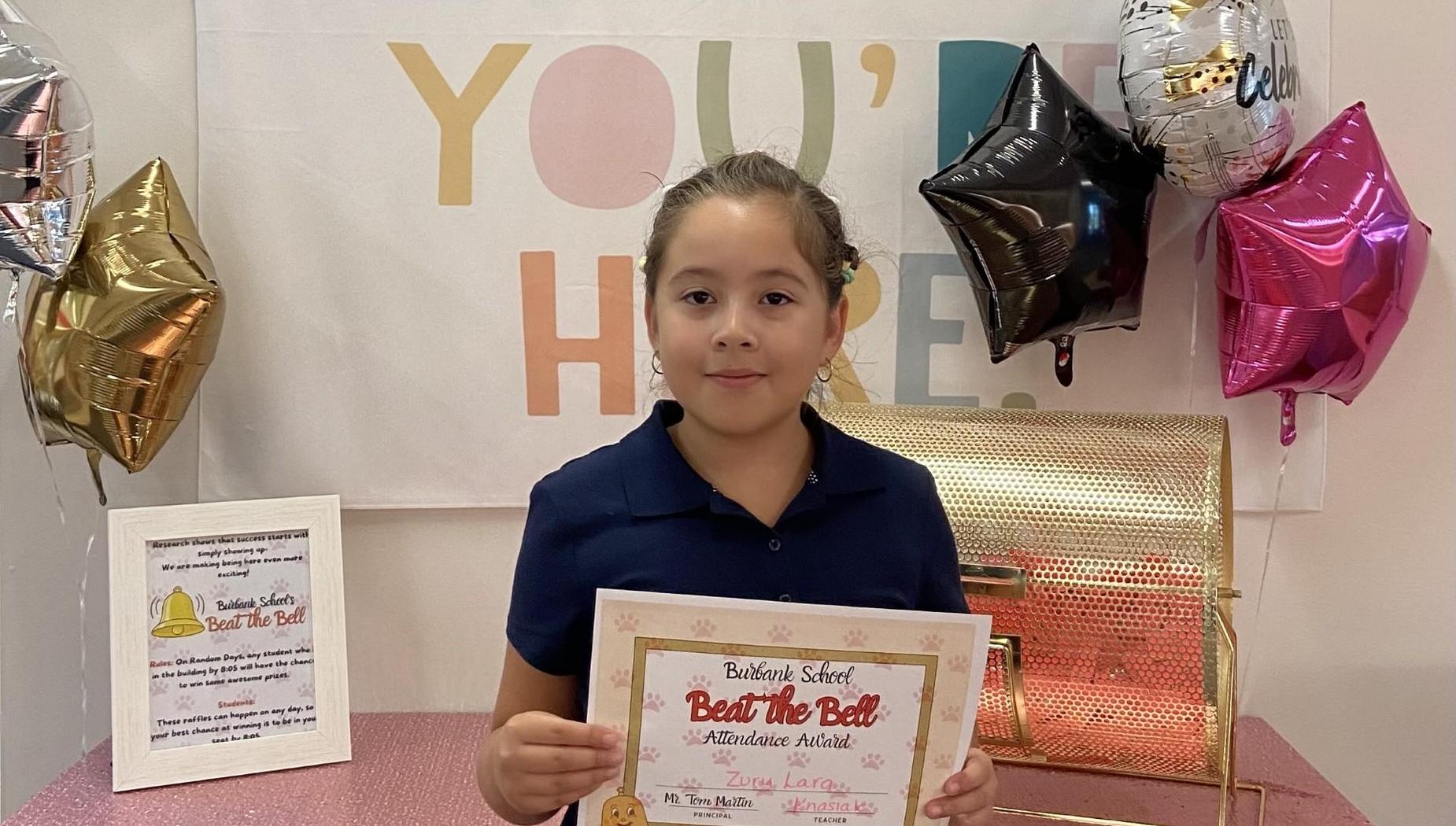 Young girl holding a certificate in front of a festive backdrop.