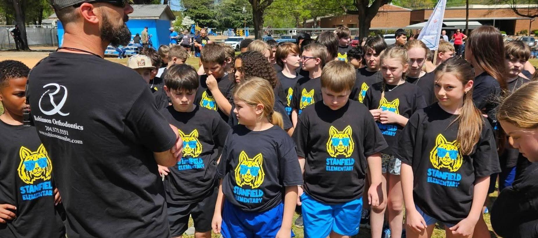 Group of students in matching shirts gathered outdoors, listening to an adult.