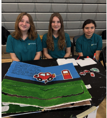Three students pose behind a project showcasing a landscape with cars and a gas station.