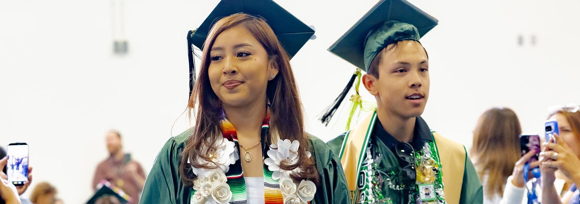 A female graduate in a green gown walks the processional aisle wearing a serape-style stole and a large white floral lei. A male graduate follows behind in a green gown with a braided green and gold lei and pins. Families watch and photograph from the sides.