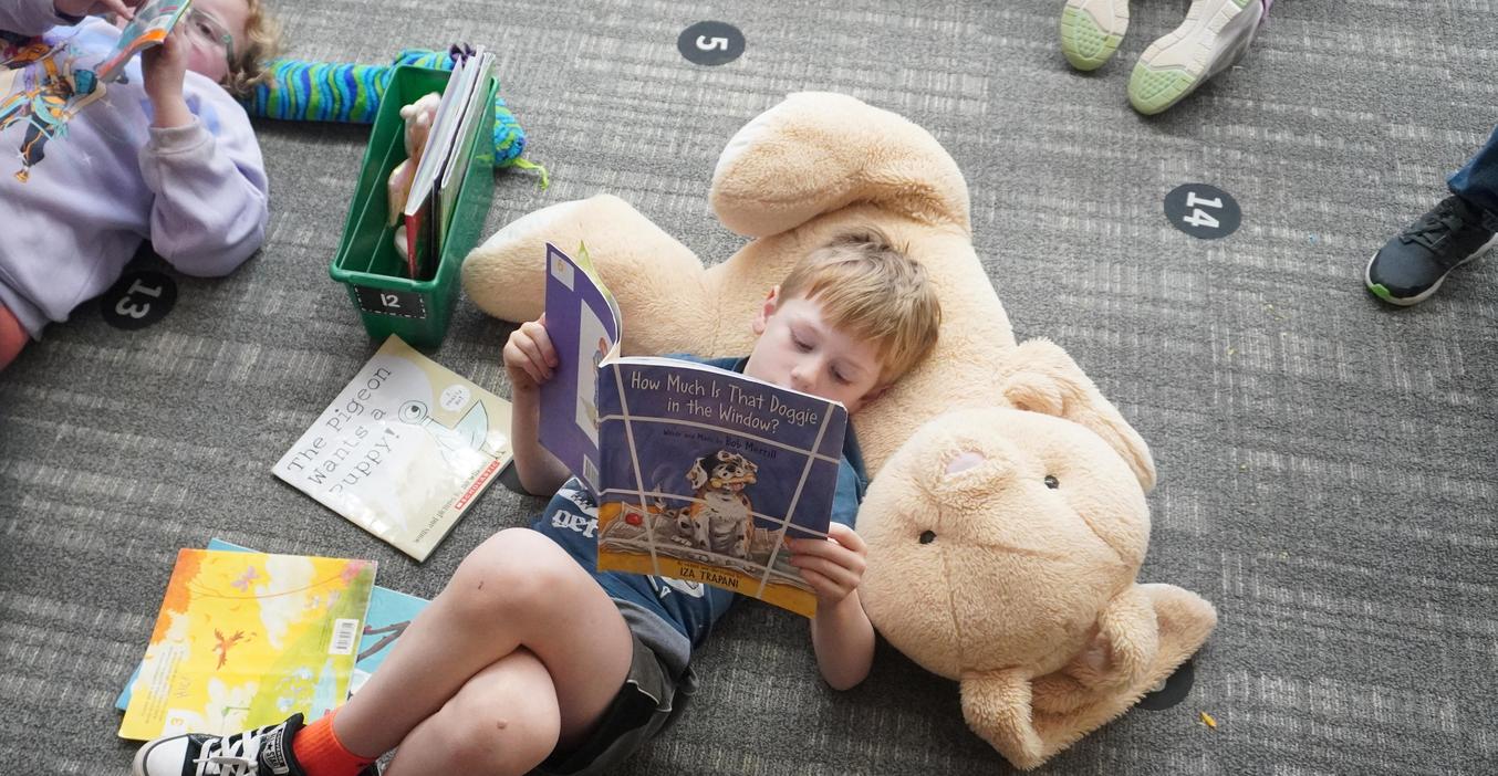 A student uses a stuffed animal as a pillow as he reads a book.