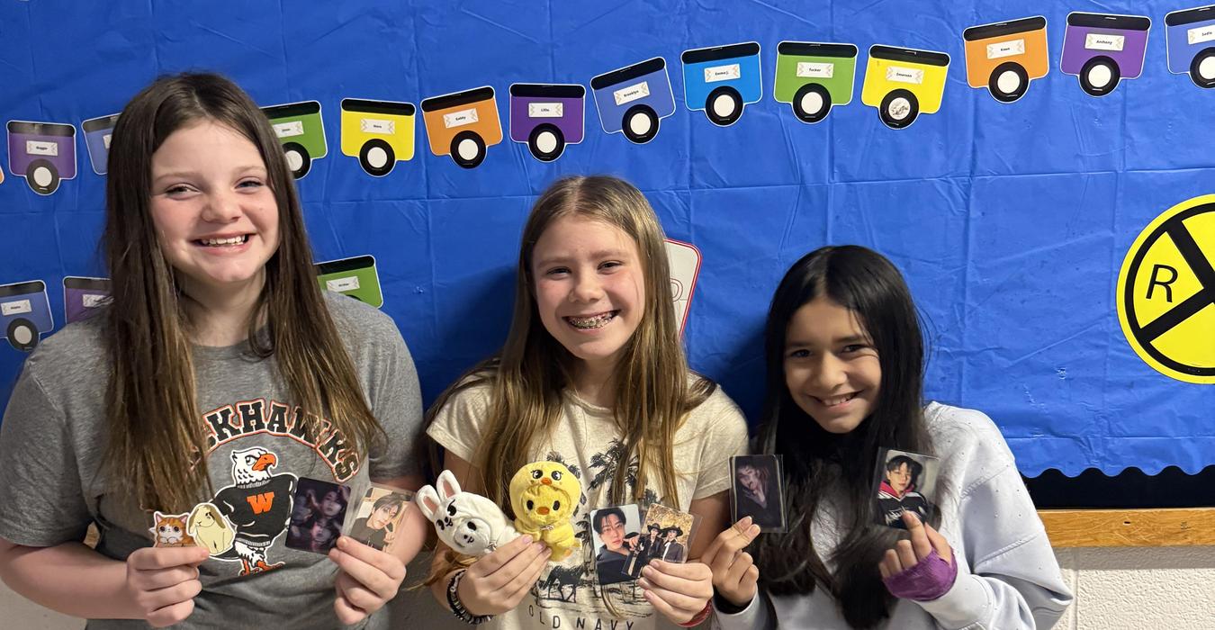 Three girls holding small photos and toys, smiling in a classroom setting.
