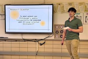Presentation with a man holding a paper by a screen showing a research question.