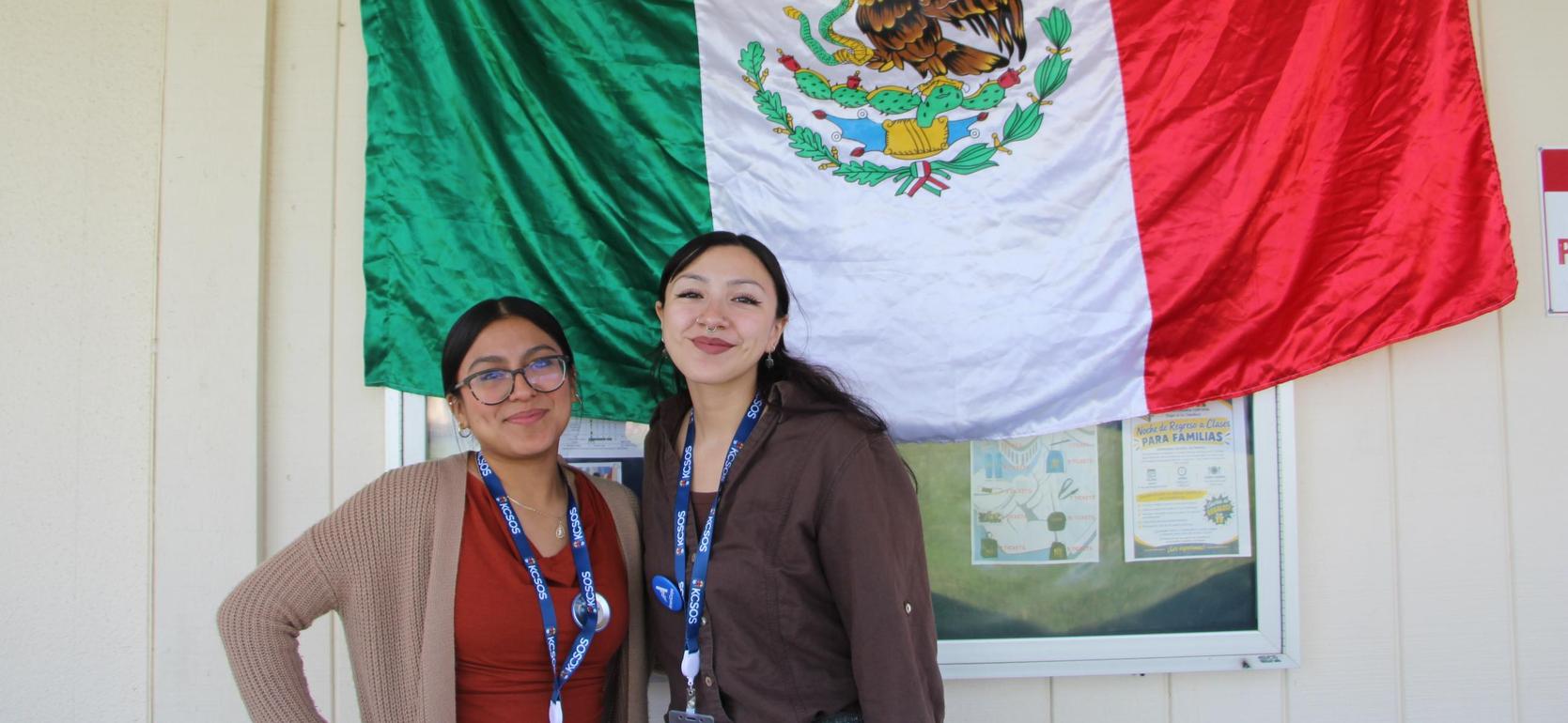 Two women smiling in front of the Mexican flag.