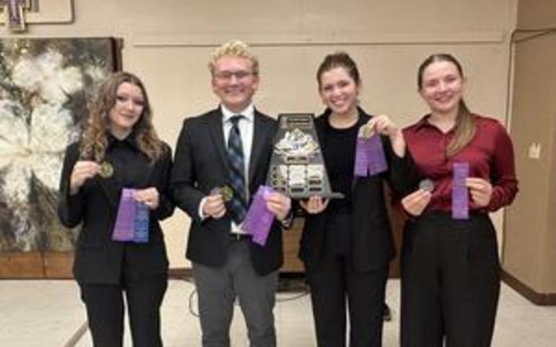 Four students in formal attire holding awards and a trophy in a classroom.