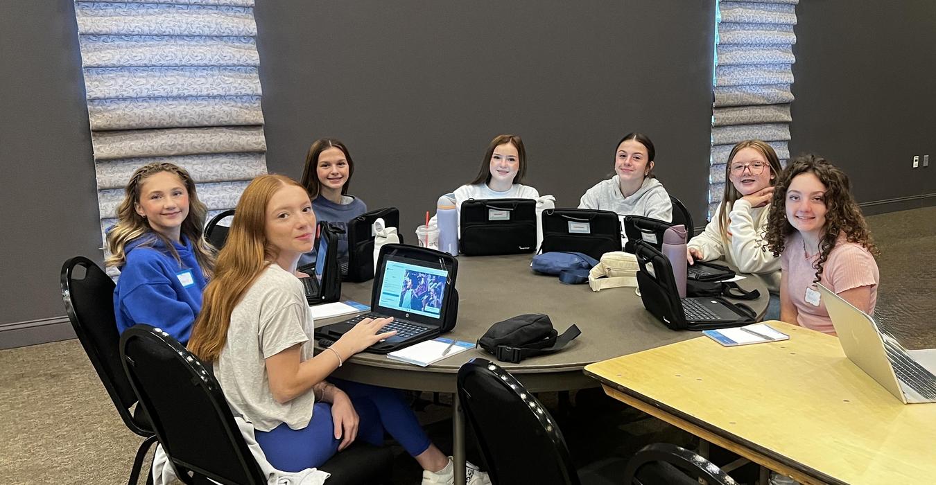 A group of seven girls sitting around a table with laptops.