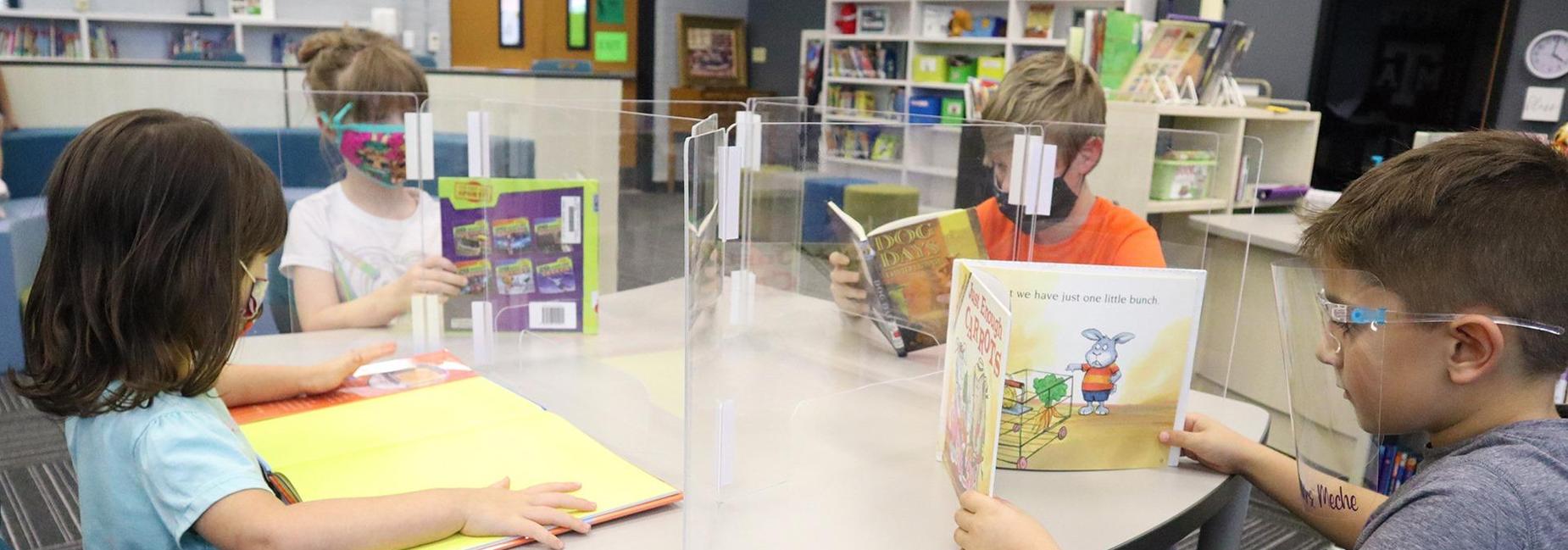 4 students reading, wearing face shields face each other with plexiglass shields between them