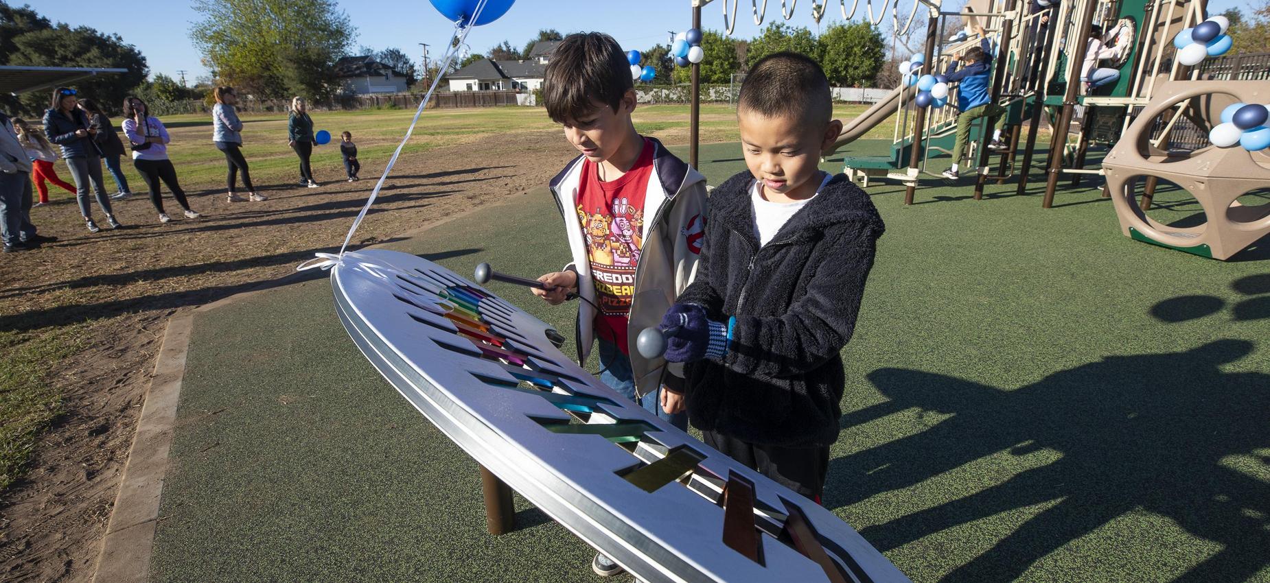 Two children playing a colorful musical instrument at a playground with people in the background.