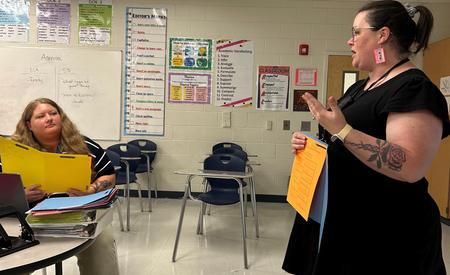 Two women in a classroom discussing with documents in hand, students seated in background.