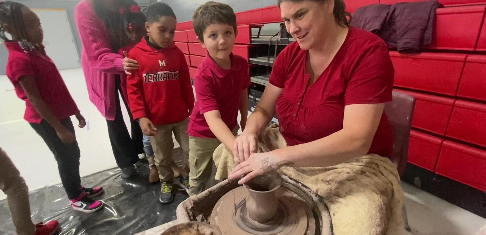 A woman guides a boy in making pottery on a wheel, surrounded by children watching.
