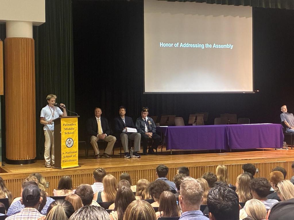 A student speaking at the podium in the auditorium with Dr. Scheibenhofer and Dr. Spering also on stage.