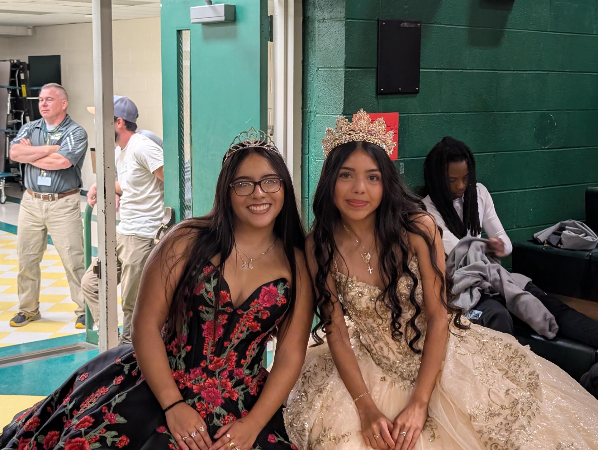 Two girls in formal dresses and crowns sitting against a wall with others in the background.