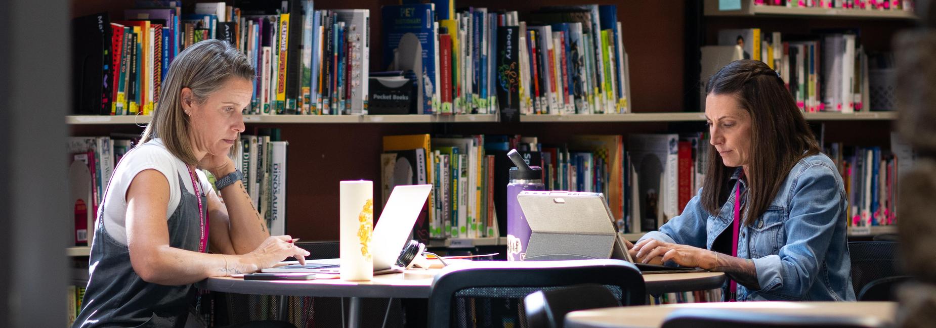 Two women working on laptops at a table in a library.