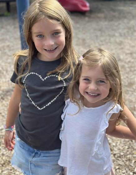 Two girls stand together, smiling on a playground with a gravel surface.