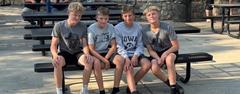 Four boys sitting together on a bench outdoors, smiling at the camera.