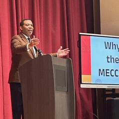 A speaker addressing an audience at a podium with a presentation screen.