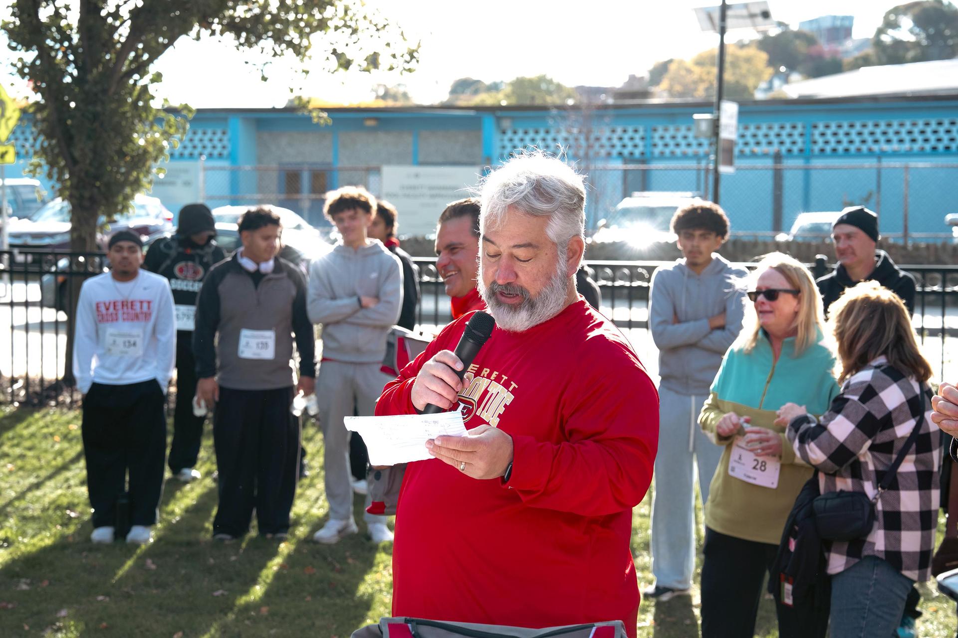 A man in a red sweatshirt speaks into a microphone while participants look on.