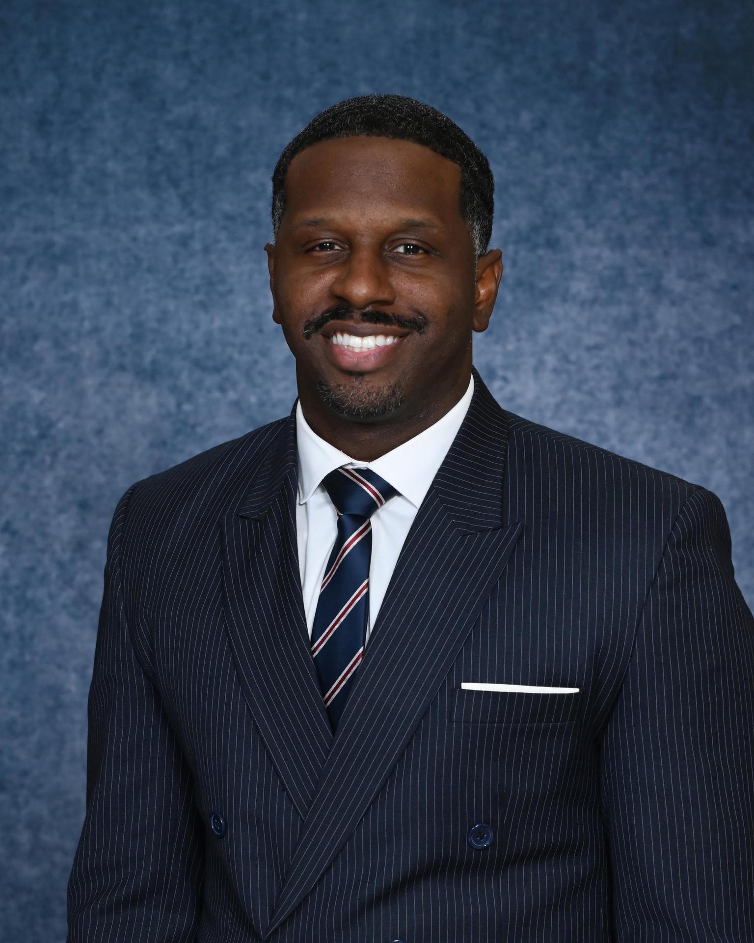 Smiling man in a navy suit and tie with a blue background behind him.