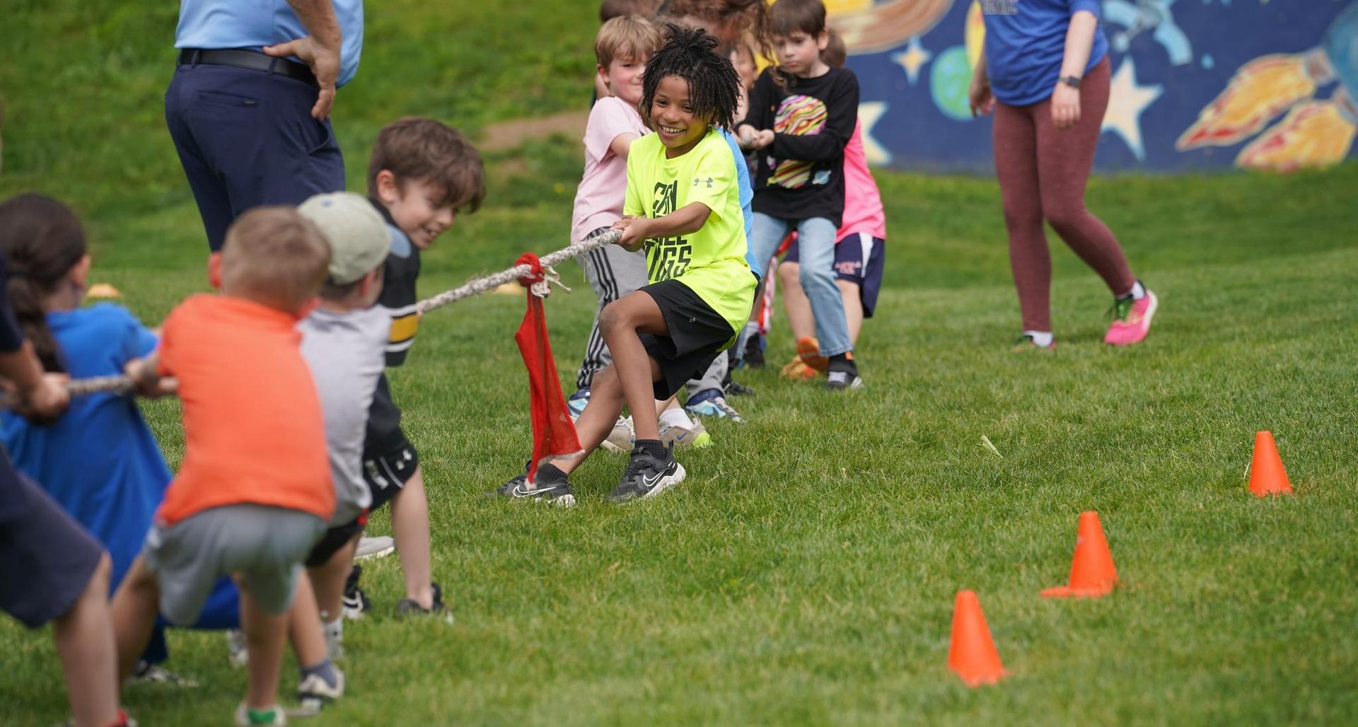 Kids engaged in a tug-of-war game on a grassy field with cones in the background.