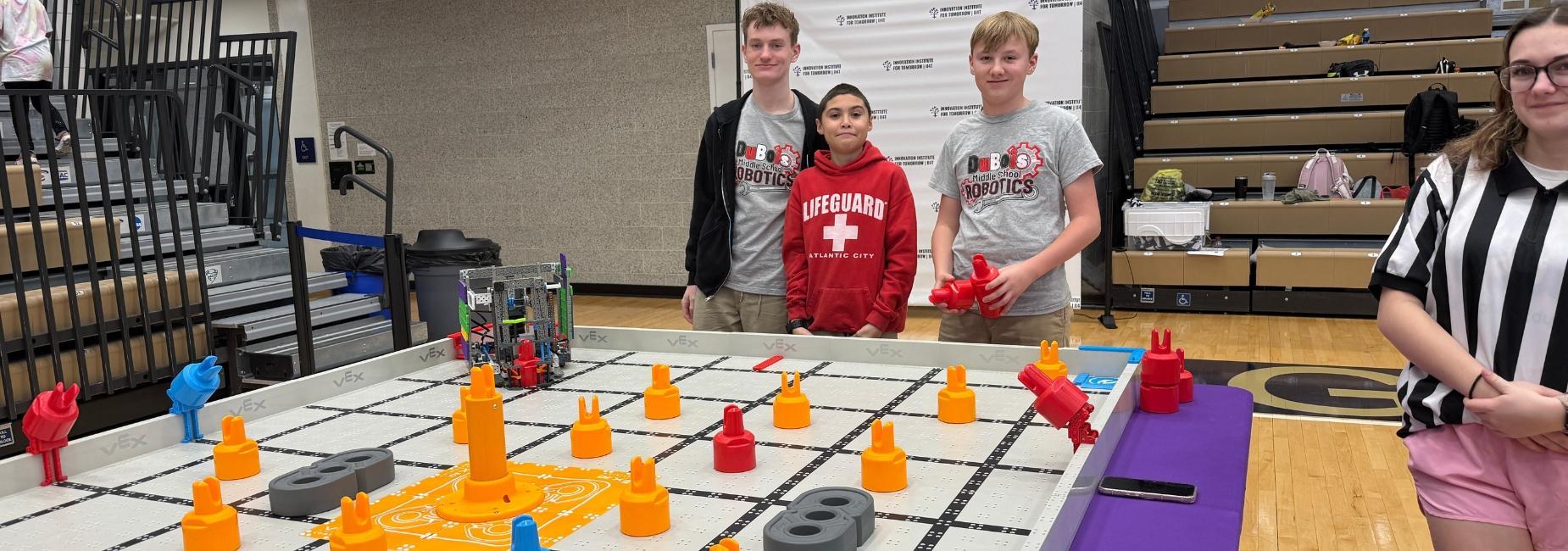 Three students stand by a colorful robotics competition board with various game pieces.