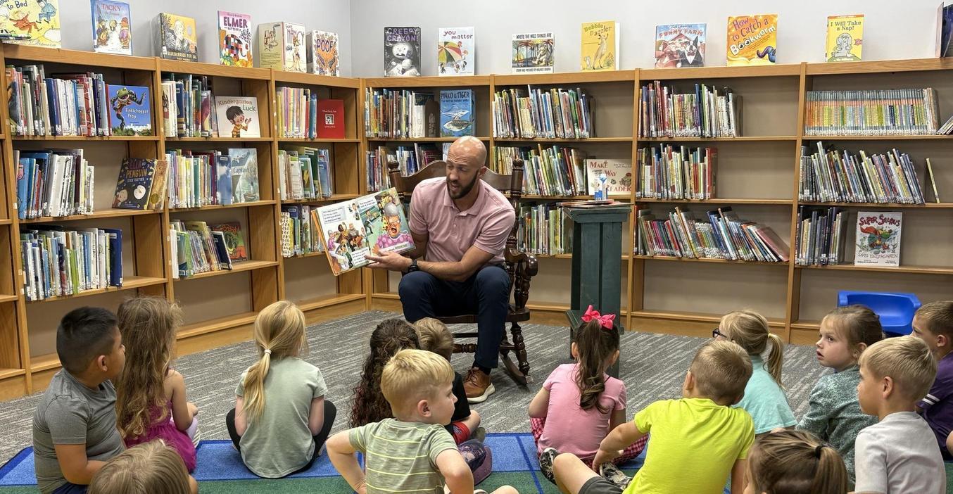 McFall Principal Sleeman reads a book to a group of children sitting on the floor in a library.
