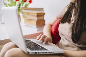 A woman sits on a chair with a laptop. Her hand rests on the touchpad of the laptop.