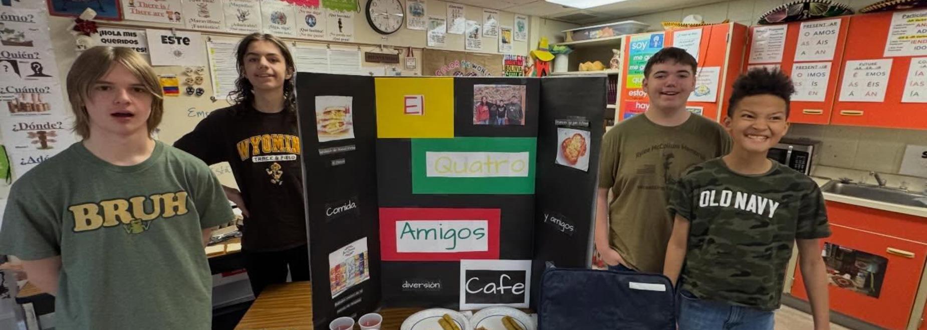 Four students stand by a display showcasing their food project titled 'El Cuatro Amigos Cafe.'