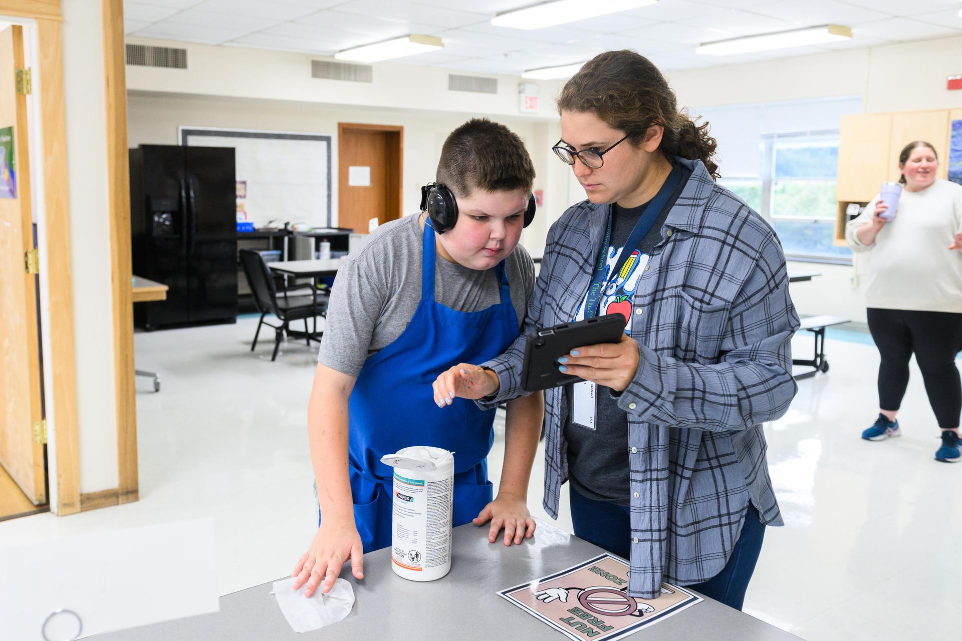 Student on right showing AAC device to student wearing headphones and blue apron on left.
