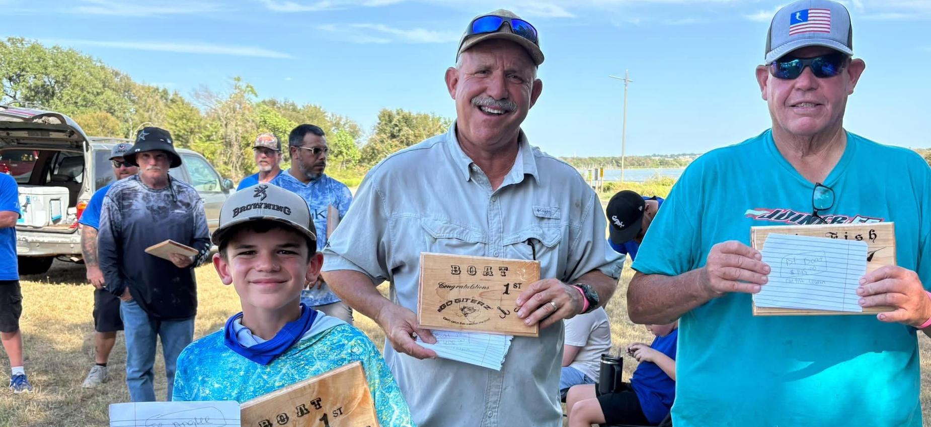 Three people holding wooden plaques, smiling in a gathering outdoors.
