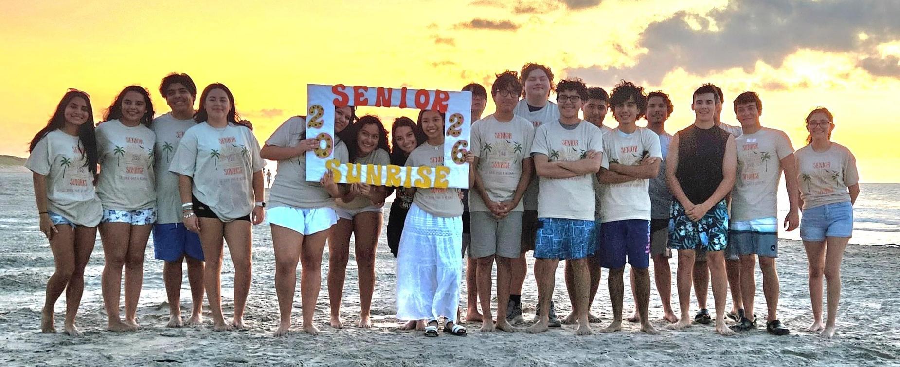 Group of graduates posing on the beach at sunrise with a banner reading 'Senior Sunrise 2023'.