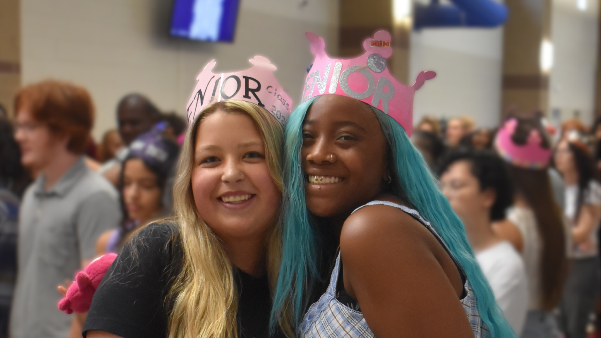 Two smiling girls in pink 'Senior' crowns at a crowded event.