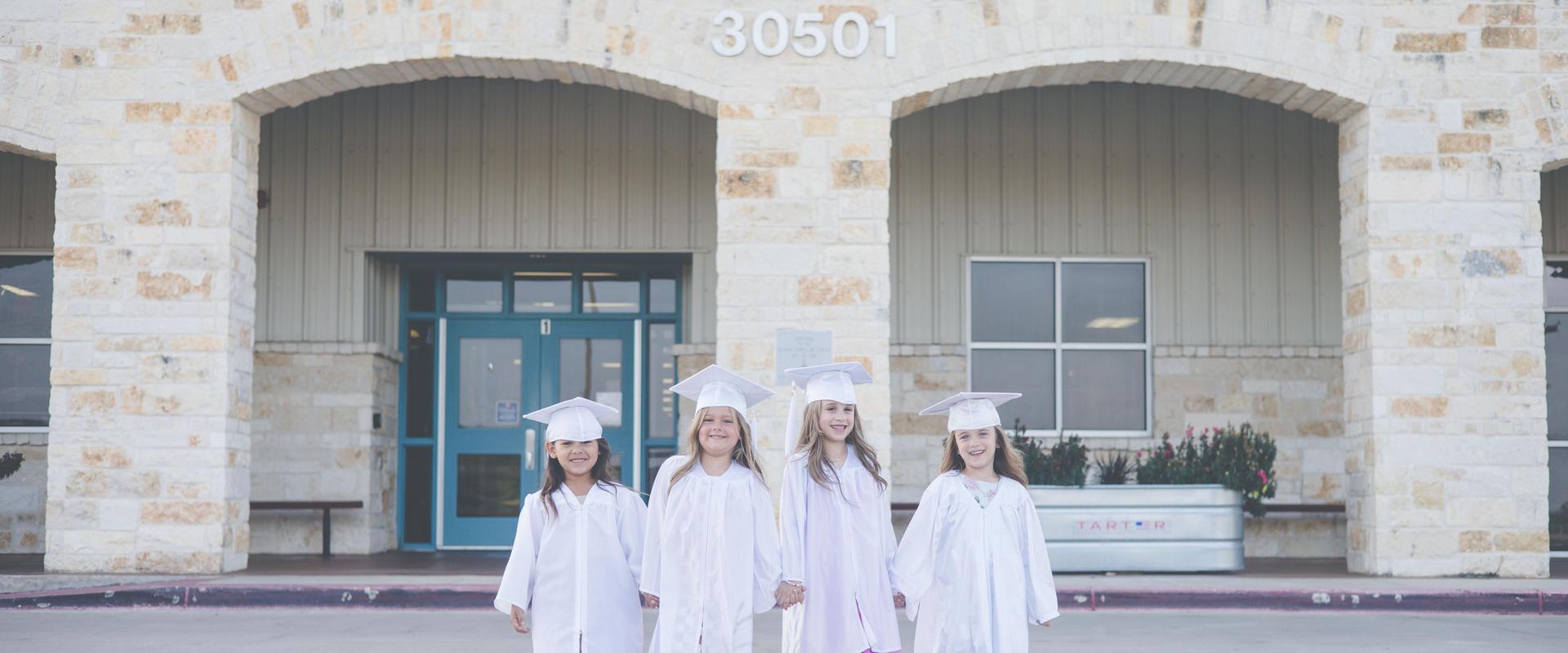 Four girls in white graduation gowns and caps stand together in front of a school building.