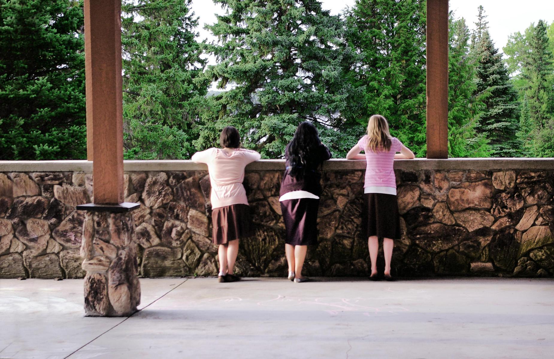 Three women lean against a stone railing, gazing at a forested landscape.