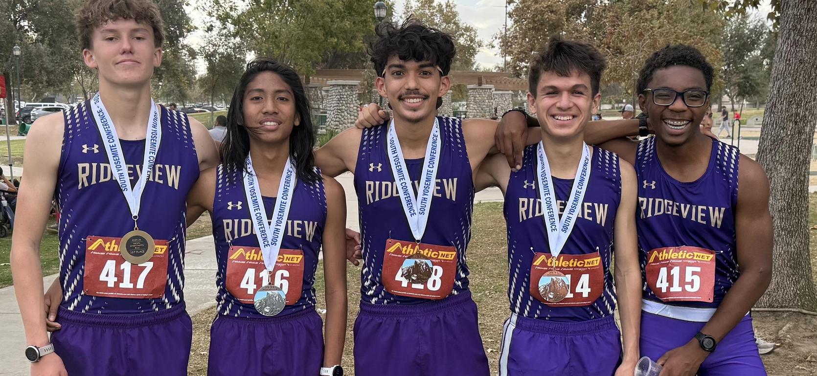 Five runners in athletic gear displaying medals at an outdoor event.