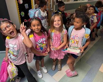 Group of smiling children in a school hallway, wearing backpacks.