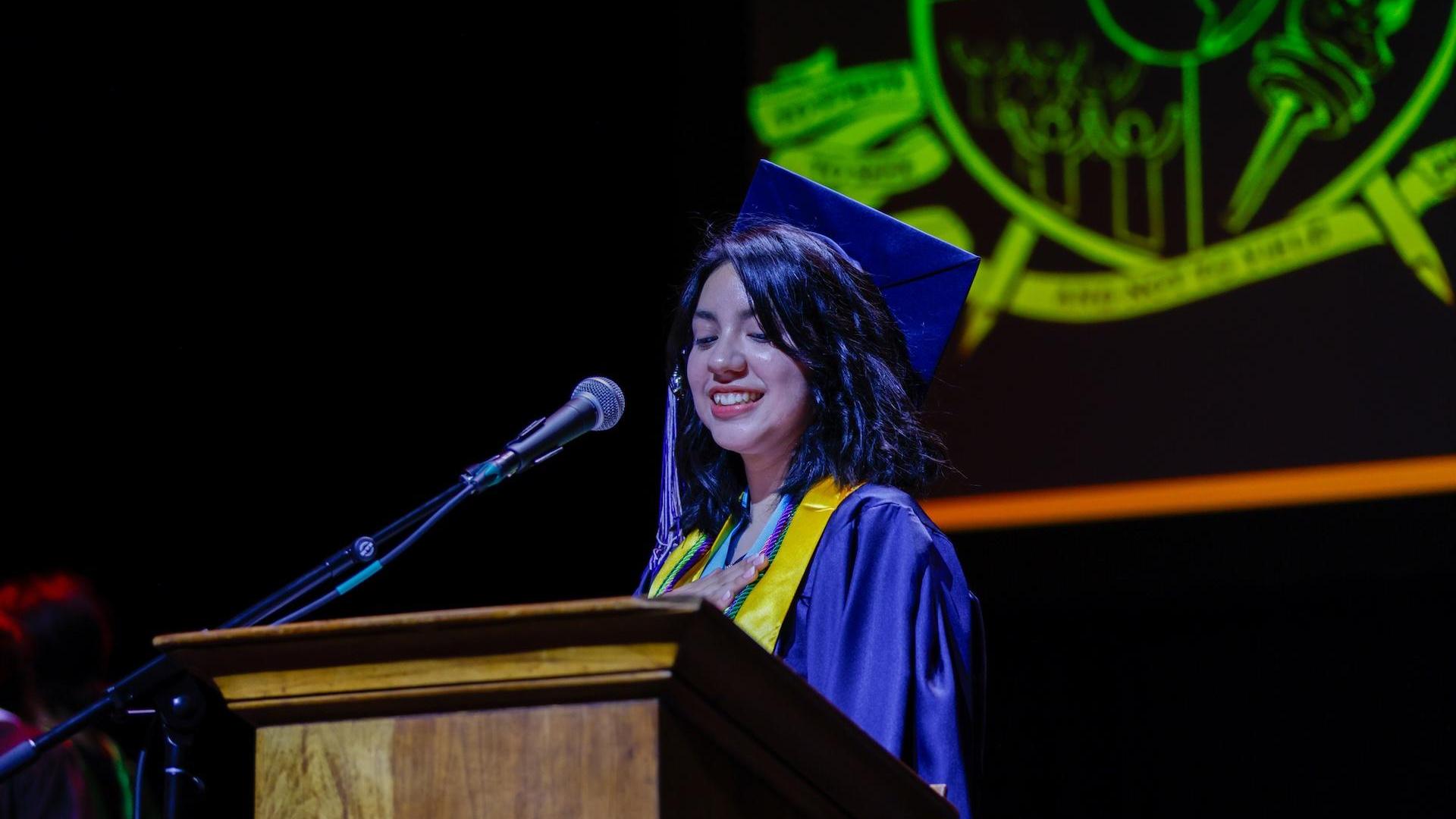 A graduate in a cap and gown smiles while speaking at a podium.
